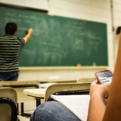 Student texting in a classroom while teacher is writing on the blackboard.